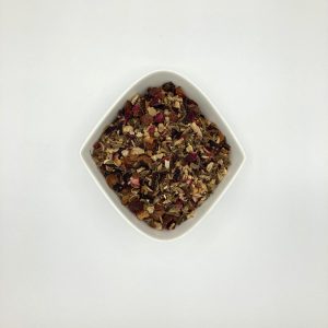 Eine weiße, rautenförmige Porzellanschale enthält rötlichbraune Kräuter- und Gewürzstückchen mit roten Hagebuttenschalen und Hibiskusblüten.